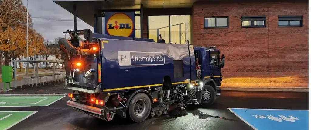 Sopmaskin från PL Utemiljö AB utför höstsopning på parkeringsyta utanför livsmedelsbutik – fokus på renhet och tillgänglighet.