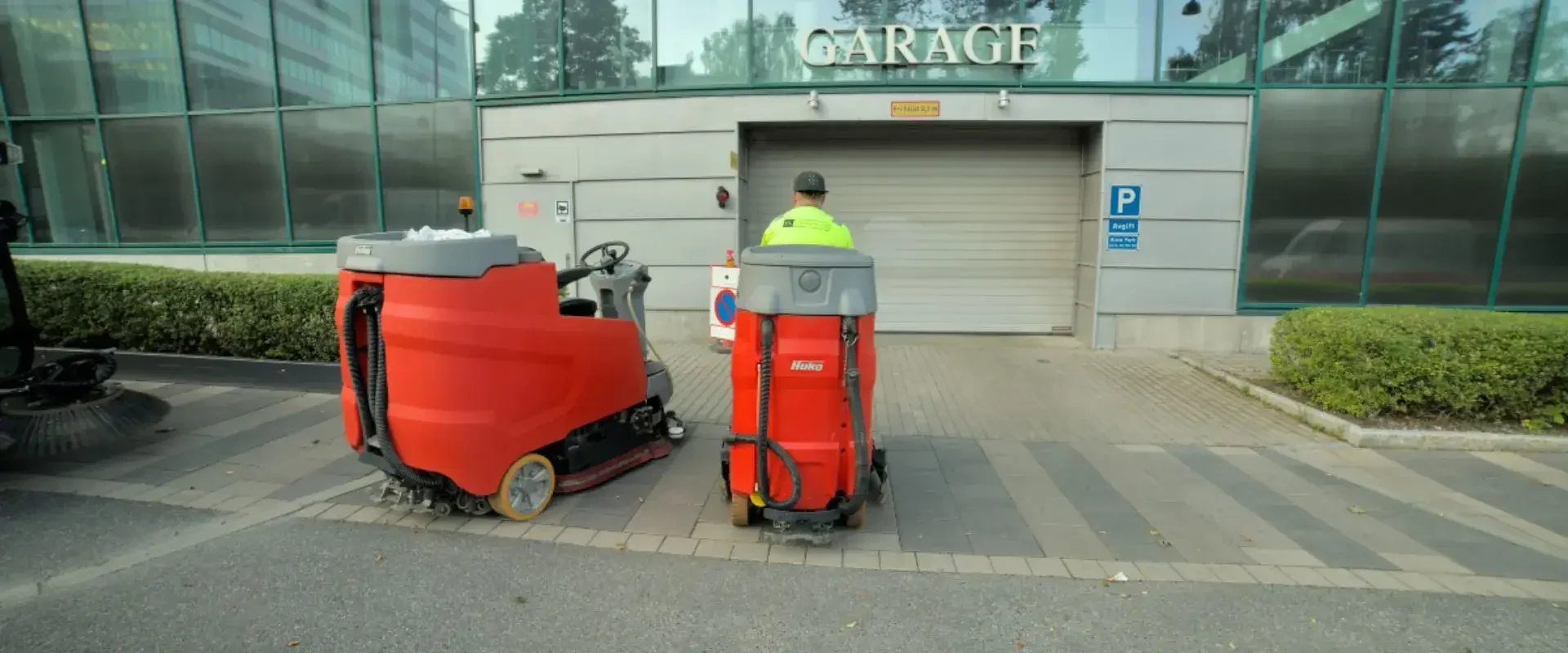 Två röda städmaskiner och en person i arbetskläder framför en garageport till ett parkeringsgarage